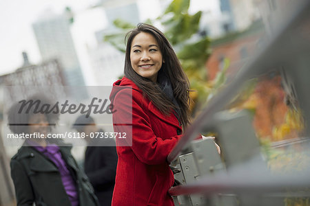 City life. A woman in a red winter coat leaning on a railing, taking time out. Two people in the background. City landscape of buildings.