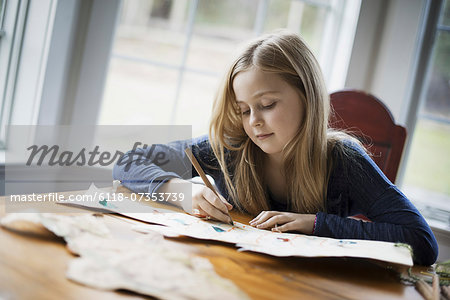 A family home. A young girl sitting at a table drawing on a large piece of paper. Holding a pencil.