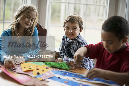 Children in a family home.  Three children creating pictures, with glue and stickers. Craftwork.