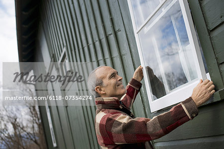 An organic farm in winter. Maintenance. A man holding a a storm window panel against the window frame.