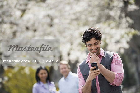People outdoors in the city in spring time. Cherry blossom on the trees. A man checking his cell phone, and two people behind him.