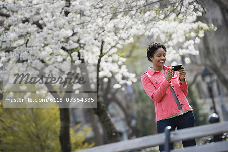 Outdoors in the city in spring time. New York City park. White blossom on the trees. A woman holding her mobile phone and smiling.