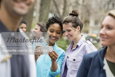 People outdoors in the city in spring time. New York City park. Two women in a group of friends, looking at a cell phone and smiling. People outdoors in the city in spring time. New York City park. Two women in a group of friends, looking at a cell phone and smiling.
