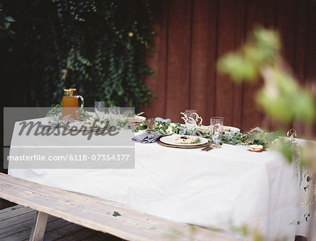 A table laid for a special meal. Place settings, with plates and cutlery. Glasses. A white table cloth and bench seat. Plates of food.