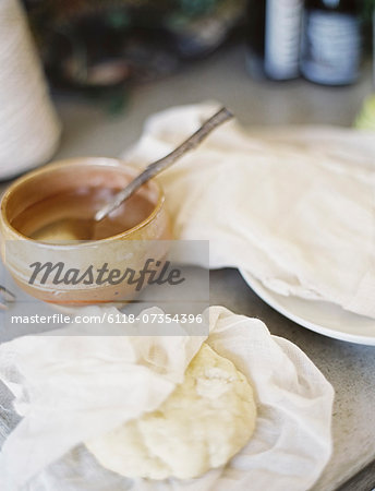 A tabletop in a domestic kitchen. A bowl of liquid with a spoon in it. A block of fresh pastry partly wrapped in a muslin square.