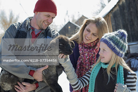 Winter scenery with snow on the ground. A man holding a young lamb, and a child stroking its chin.
