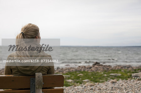 A woman with long blonde hair wearing a brown hooded coat, seated on a bench looking out to sea.