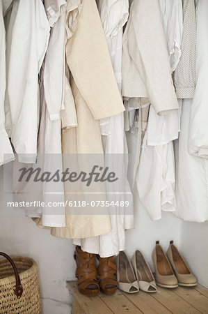 A closet with cream and white clothes, jackets, shirts and tunics hanging up. A row of women's shoes arranged neatly on the floor.