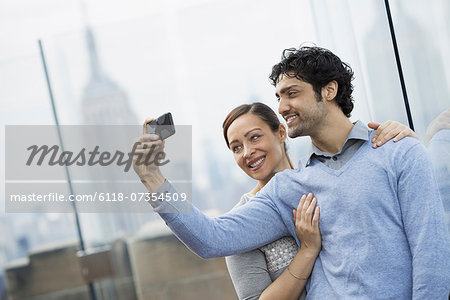 New York City. An observation deck overlooking the Empire State Building. A young couple taking photographs with a mobile phone.
