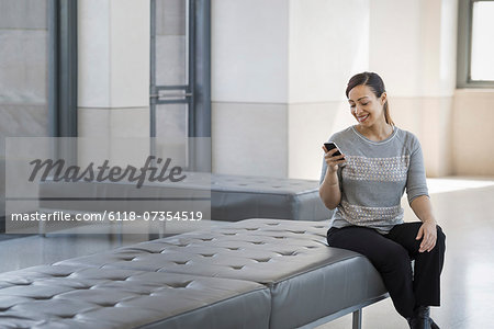 Urban Lifestyle. A young woman sitting on a seat in a building, using her mobile phone.