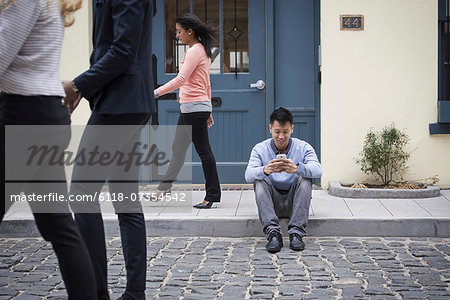 Young people outdoors on the city streets in springtime. A man sitting on the ground checking his phone, and three passers-by.
