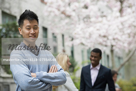 City life in spring. Young people outdoors in a city park. A man with his arms folded and two people talking in the background under the trees in blossom.