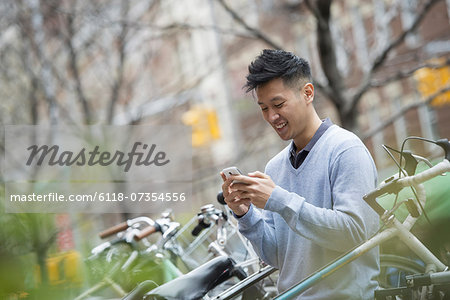 City life in spring. A man in a blue sweater by a row of parked bicycles. Checking his messages on a smart phone.