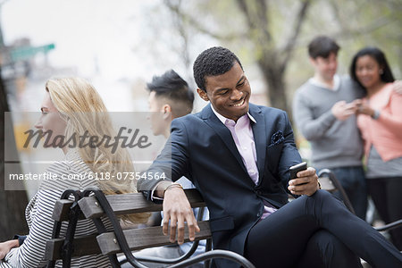 City life in spring. Young people outdoors in a city park. A group of men and women gathered around a park bench. Two checking a smart phone for messages. City life in spring. Young people outdoors in a city park. A group of men and women gathered around a park bench. Two checking a smart phone for messages.