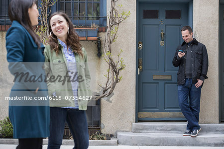 Two women talking as they walk down a street. A man leaning against a house door checking his phone.