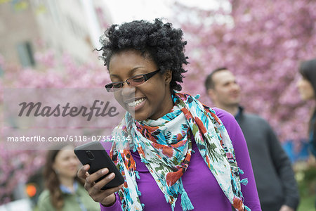 A group of people under the cherry blossom trees in the park. A young woman smiling and checking her phone.