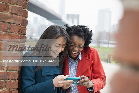 New York city, the Brooklyn Bridge crossing over the East River. Two women, side by side, checking the screen of a smart phone, and laughing.