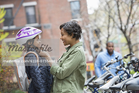 A New York city park in the spring. A boy in a cycle helmet, being fastened by his mother, beside a bicycle rack.