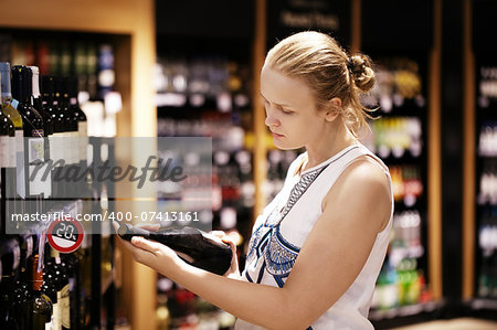 Woman shopping for wine or other alcohol in a bottle store standing in front of shelves full of bottles and holding bottle in her hand and reading inscription
