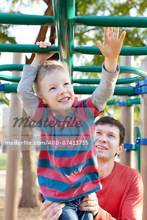 cheerful little boy playing at monkey bars and his father helping him at the playground