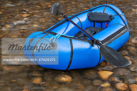 a blue pacraft (one-person light raft used for expedition or adventure racing) with a kayak paddle against a shallow river