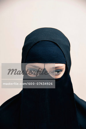 Close-up portrait of young woman wearing black, muslim hijab and muslim dress, eyes looking to the side showing eye makeup, studio shot on white background