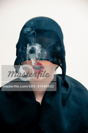 Close-up portrait of young woman wearing black, muslim dress and black, hijab covering part of head, while blowing smoke rings from red lips, studio shot on white background