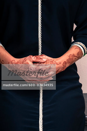 Woman sitting indoors with close-up of folded hands painted with henna in arabic style, wearing a typical black, arabic, muslim dress, studio shot