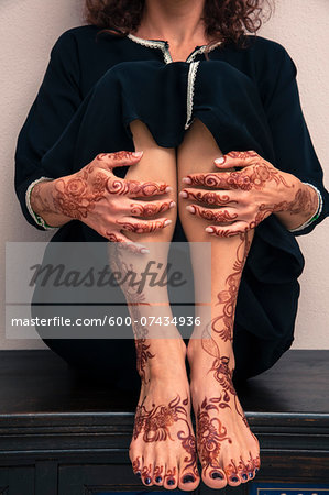 Low section of woman sitting indoors, showing legs, feet and hands painted with henna in arabic style, wearing a typical black, arabic, muslim dress, studio shot