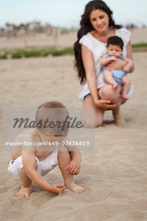 Toddler digging in sand with hand, mother and baby in background