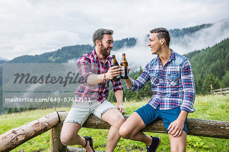 Two male friends drinking beer on fence, Tyrol Austria