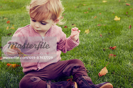 Toddler sitting on grass looking at autumn leaf