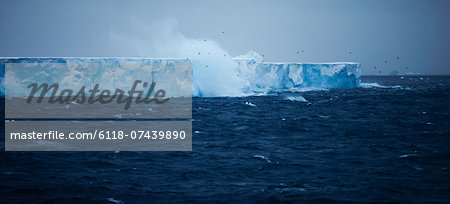 A large iceberg with steep sides, floating on the water. Waves and spray rising. Calving event.