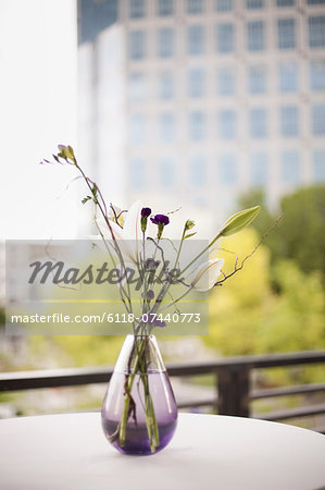A table on a terrace in the city. A vase of flowers. Small purple flowers, and white lily and orchid blooms.