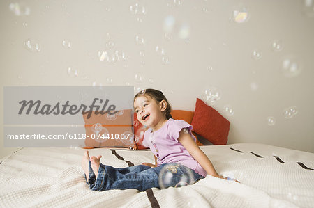 A young girl with brown eyes and dark hair in bunches sitting on a bed laughing. Bubbles floating in the air.