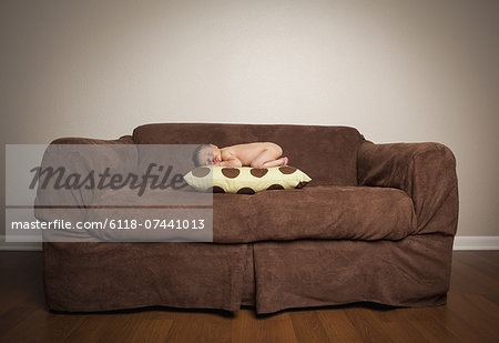 A naked newborn baby lying on his front, sleeping on a pillow on a brown couch.