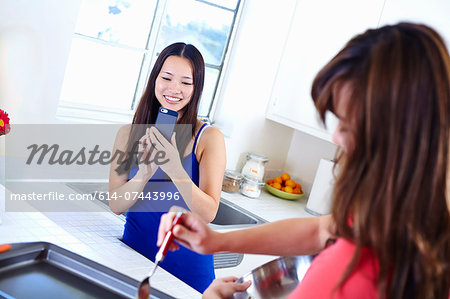 Two young women in kitchen taking photograph with mobile phone