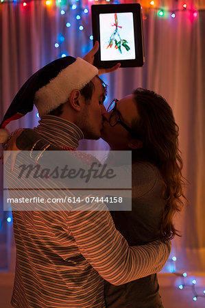 Young couple kissing whilst holding up digital tablet at christmas