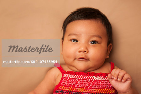 Close-up portrait of Asian baby lying on back, wearing red dress, looking at camera and smiling, studio shot on brown background