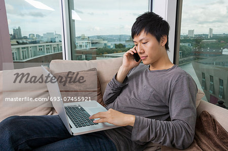 Asian mid adult man on call while using laptop in living room
