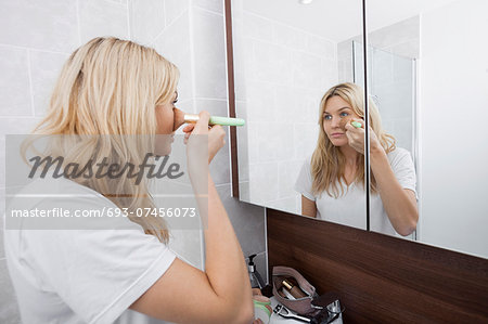 Young woman applying blush while looking at mirror in bathroom