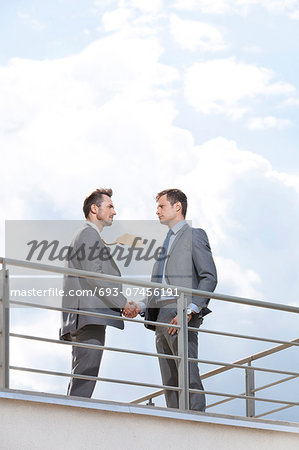 Side view of businessmen shaking hands at terrace railings against sky