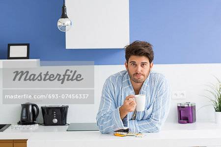 Portrait of young ill man holding coffee mug while leaning on kitchen counter