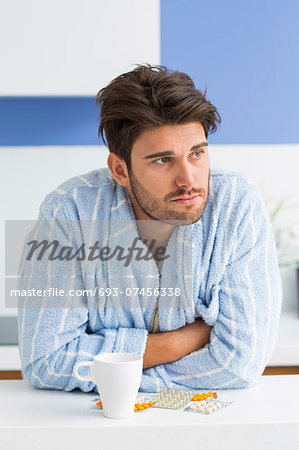 Young ill man with coffee mug and medicine leaning on kitchen counter
