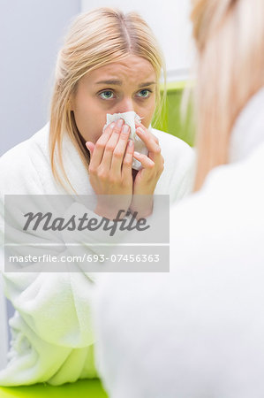 Young woman blowing nose in tissue paper while looking at mirror