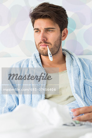 Sick man with thermometer in mouth sitting on bed