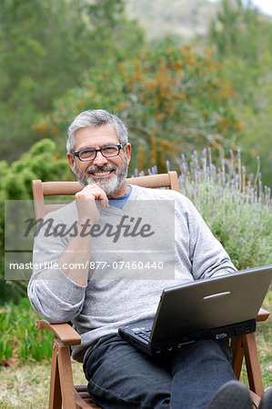 Man sitting in his garden with laptop, smiling and wearing glasses