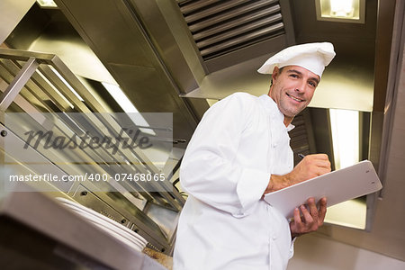 Low angle portrait of a smiling male cook writing on clipboard in the kitchen