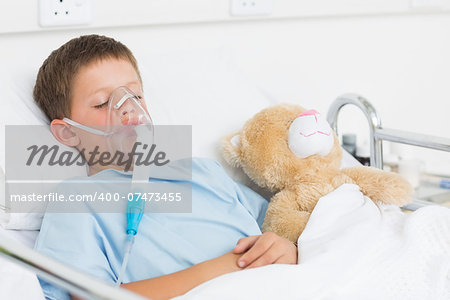 Sick boy wearing oxygen mask sleeping beside teddy bear in hospital bed
