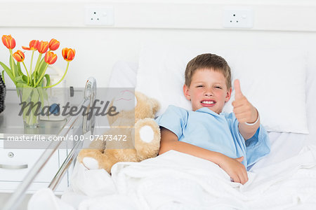 Portrait of little boy gesturing thumbs up with teddy bear in hospital bed